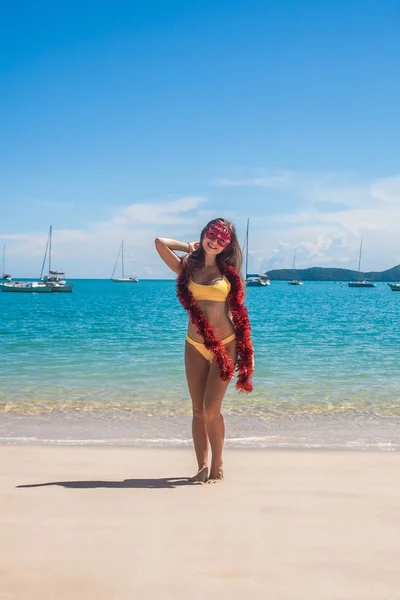 Młoda kobieta w bikini nosić śmieszne Wesołych Świąt Bożego Narodzenia okulary i czerwony blichtr stojący na plaży — Zdjęcie stockowe