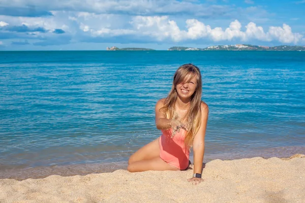 Uśmiechnięta młoda kobieta w strój kąpielowy różowy, rzucanie piasku na tropikalnej plaży — Zdjęcie stockowe