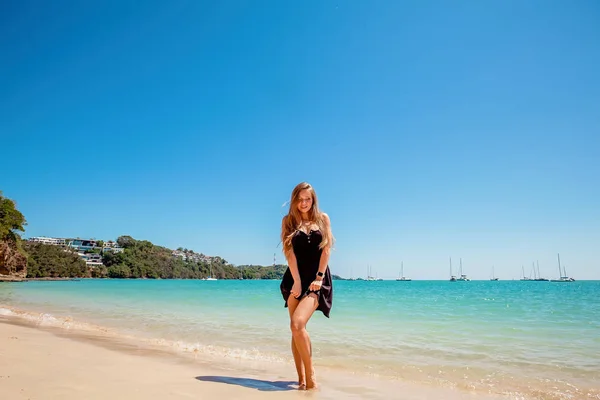 Vacker ung kvinna med långt hår i en svart klänning stående på sand nära havet — Stockfoto