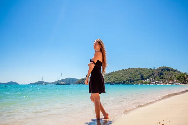 Mooie jonge vrouw met lang haar in een zwarte jurk staande op zand in de buurt van de zee — Stockfoto