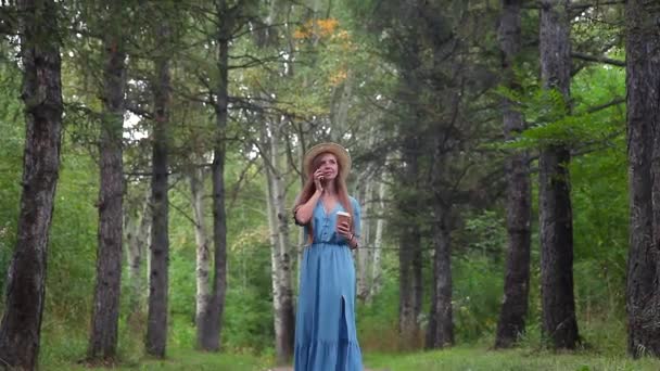 Mujer joven caminando en un bosque hablando por teléfono móvil, sosteniendo una taza de café — Vídeos de Stock