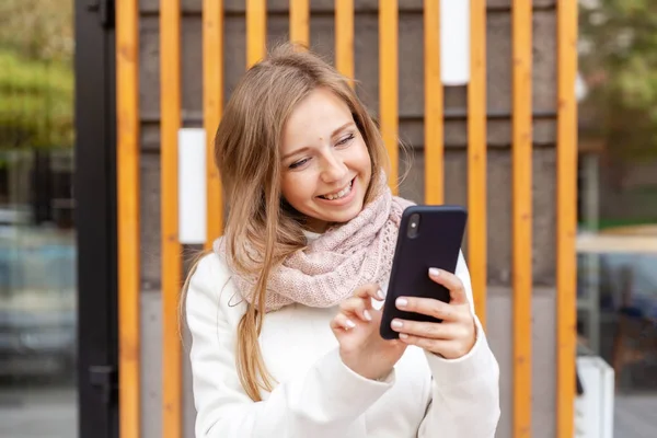 Portrait de jeune femme souriante en manteau blanc avec écharpe touchant le smartphone — Photo