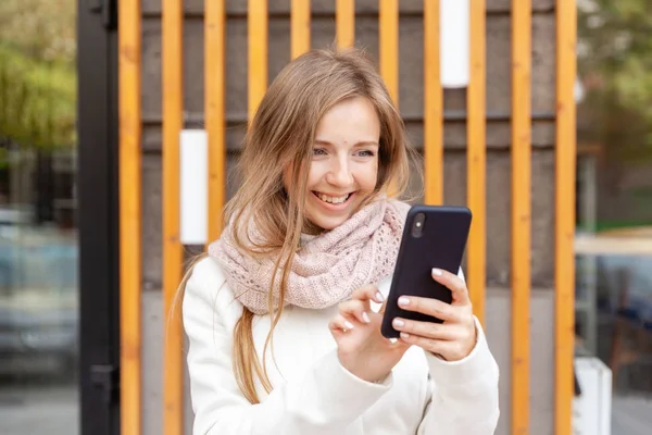 Jeune femme d'affaires souriante en manteau blanc avec écharpe travaillant avec smartphone — Photo