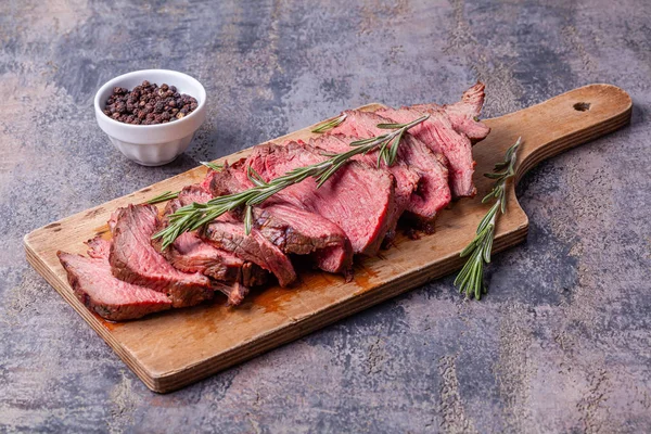 Slices of medium rare roast beef meat on wooden cutting board, pepper, rosemary — Stock Photo, Image