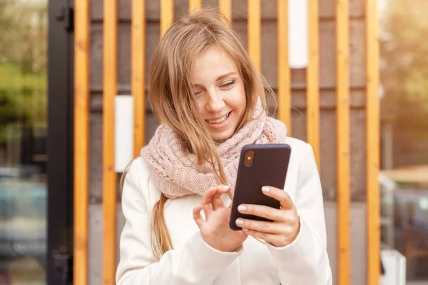 Joyeux jeune femme souriante en manteau avec écharpe en utilisant le smartphone dans les rayons de soleil — Photo