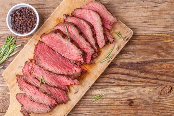 Sliced medium rare roast beef meat on wooden cutting board, pepper and rosemary