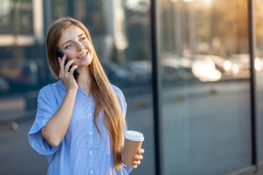 Akıllı telefondan konuşan, elinde kahve fincanı tutan mutlu ve çekici genç bir kadın.