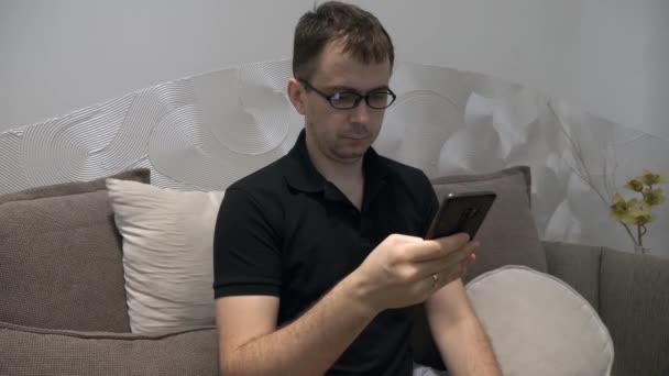 Hombre serio con camiseta de polo negro y gafas sentado en el sofá con teléfono móvil — Vídeo de stock