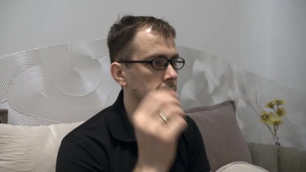 Уставший серьезный человек в футболке с черным поло сидит на диване, снимает очки — стоковое видео
