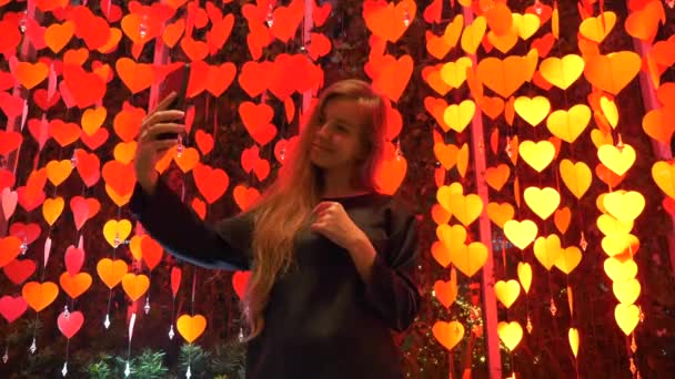Девушка блоггер с мобильным телефоном среди украшения с красной висячей бумаги сердца — стоковое видео