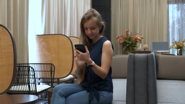 Щаслива красива молода дівчина сидить на дивані і використовує мобільний телефон — стокове відео