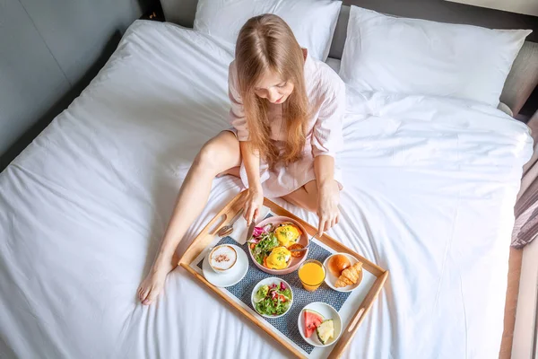 Jonge glimlachende mooie vrouw ontbijten in bed in gezellige hotelkamer — Stockfoto