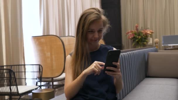 Felice bella ragazza seduta sul divano e utilizzando il telefono cellulare — Video Stock