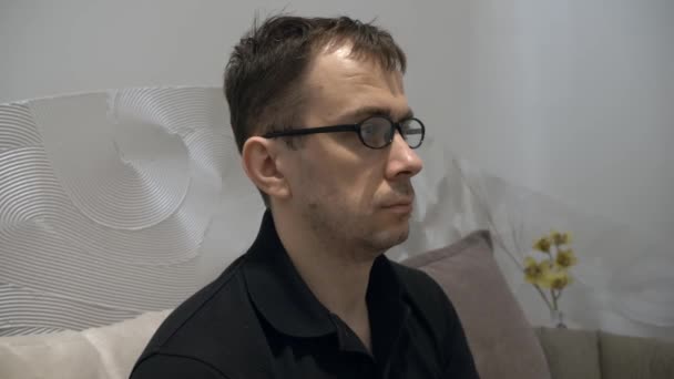 Hombre serio cansado en camiseta de polo negro sentado en el sofá, se quita las gafas — Vídeo de stock