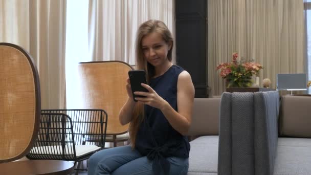 Feliz hermosa joven sentada en el sofá y usando el teléfono móvil — Vídeo de stock