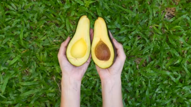Vrouw handen vasthouden en open twee gesneden helften van verse groene avocado op groen gras — Stockvideo