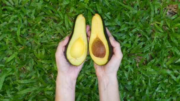 Frauenhände halten und öffnen zwei geschnittene Hälften frischer grüner Avocado auf grünem Gras — Stockvideo