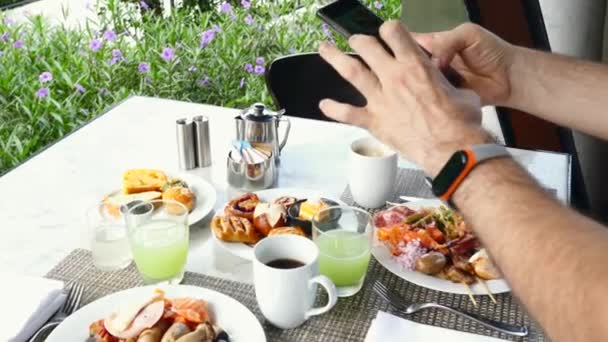 携帯電話を手にした男が写真を撮り、ホテルでビデオ朝食を作る — ストック動画