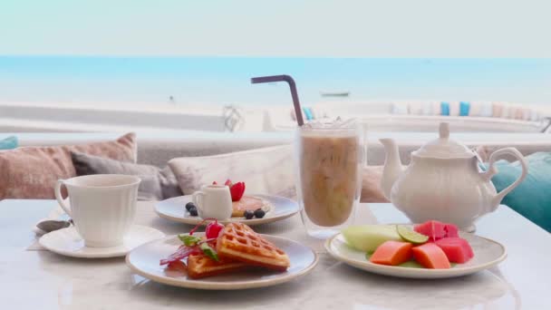 Завтрак на столе с видом на море, ледяной кофе, чайник, бельгийские вафли, блины — стоковое видео