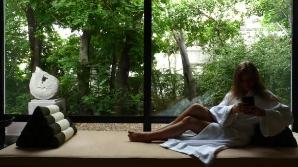 Молода жінка з мобільним телефоном лежачи на веранді біля вікна з зеленицею — стокове відео