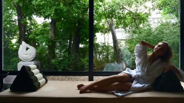 Attrayant jeune femme en peignoir blanc repose par la fenêtre. Repos fille sur canapé. — Video