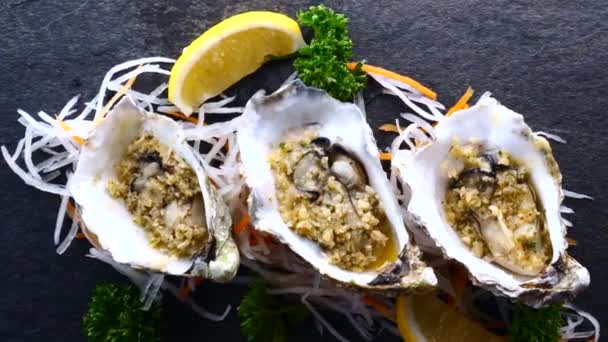 Austern in Schalen gebraten mit Knoblauch und Zitrone auf schwarzem strukturiertem Schiefer Hintergrund — Stockvideo