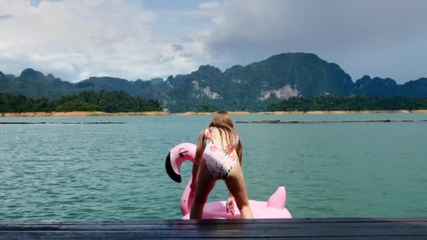 Mulher nova engraçada na roupa de banho que senta-se no Flamingo inflável rosa no lago azul — Vídeo de Stock