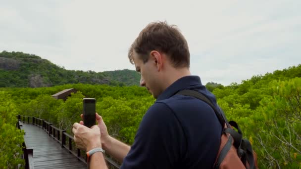 Человек с рюкзаком снимает видео в зеленом лесу, вид сзади — стоковое видео