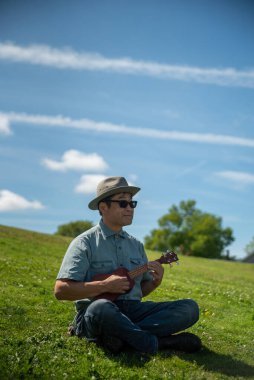 Parktaki tepede oturmuş ukulele çalan Asyalı bir adam. Vancouver BC Kanada