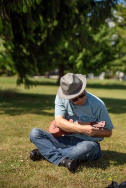 Parkta oturan ve ukulelesini ayarlayan Asyalı bir adam. Vancouver BC Kanada
