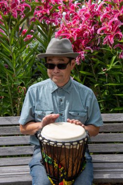 Asyalı bir adam bankta oturmuş djembe çalıyor. Vancouver BC Kanada