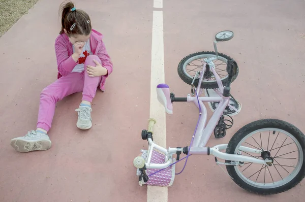 Child girl crying after bike accident. Pink female bike on floor with training wheels