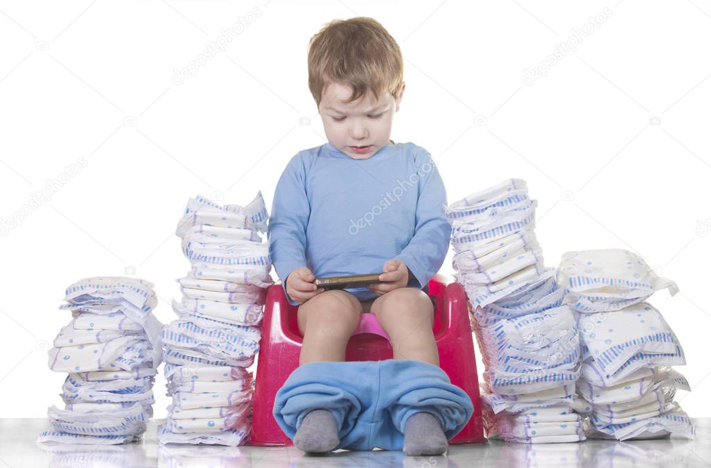 Baby boy sitting on chamber pot with smartphone and toilet paper rolls around. Potty training concept