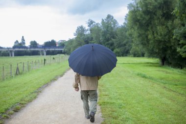 A retired man, protected by an umbrella, walks through a green meadow. Concept of financial protection through retirement pensions.
