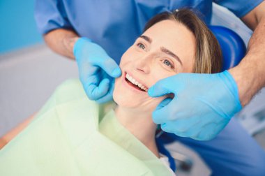 Diş hekimi muayenehanesinde bir hastanın dişlerini diş teçhizatı kullanarak muayene ediyor. Stomatoloji ve sağlık hizmetleri kavramı. Tek kullanımlık tıbbi maskeli yakışıklı erkek doktor, gülümseyen mutlu kadın..