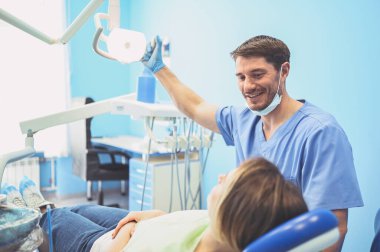 Diş hekimi muayenehanesinde bir hastanın dişlerini diş teçhizatı kullanarak muayene ediyor. Stomatoloji ve sağlık hizmetleri kavramı. Tek kullanımlık tıbbi maskeli yakışıklı erkek doktor, gülümseyen mutlu kadın..