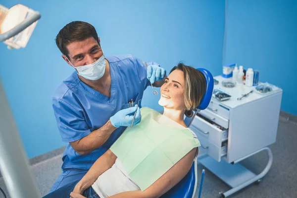 歯科室で歯科機器を用いて患者の歯を調べる歯科医 血液学と健康管理の概念 若いですハンサムな男性医師で使い捨て医療顔マスク 笑顔幸せな女性 — ストック写真