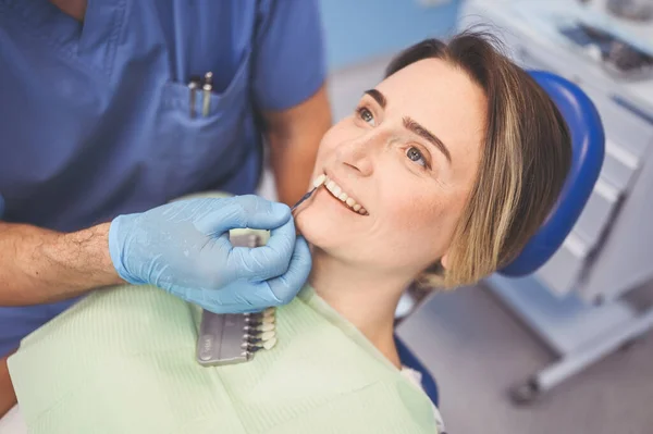 Dentist checks the level of patient\'s teeth whitening with a dentist\'s color. Dental equipment in dentistry office. Stomatology concept. Doctor\'s hands in medical gloves and smiling happy woman.