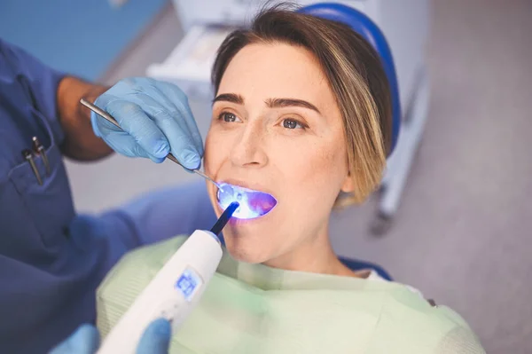 Dentist doctor using dental curing light equipment for filling, after examining a patient\'s teeth in dentistry office. Stomatology and health care concept. Close up young happy woman in dental chair