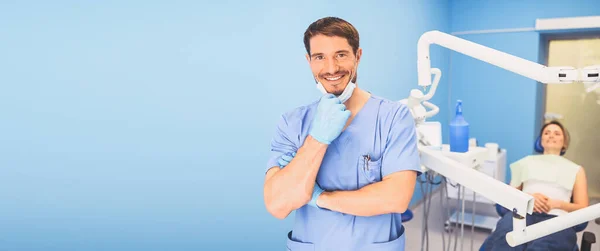 Mavi üniformalı, tek kullanımlık tıbbi maskeli, dişçi muayenehanesinde ekipmanı olan ve dişçi koltuğunda mutlu bir hastası olan genç, gülümseyen erkek doktor. Stomatoloji konsepti. Boşluğu kopyala