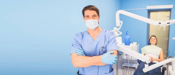 青い医療制服を着た若い笑顔ハンサムな男性医師 歯科事務所の機器と歯科椅子の幸せな患者と使い捨ての医療面マスク ストーモロジーの概念 スペースのコピー — ストック写真
