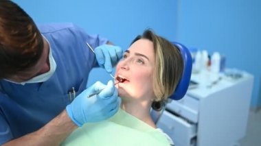 Diş hekimi muayenehanesinde bir hastanın dişlerini diş malzemesi kaşığıyla muayene ediyor. Stomatoloji ve sağlık hizmetleri kavramı. Tek kullanımlık tıbbi maskeli doktor, gülümseyen mutlu kadın..