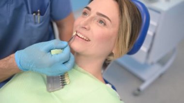 Diş hekimi hastanın diş beyazlatma seviyesini dişçinin rengiyle kontrol ediyor. Diş hekimliğinde dişçi aletleri. Stomatoloji konsepti. Eldivenli doktor elleri ve gülümseyen mutlu kadın..
