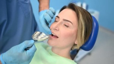 Diş hekimi muayenehanesinde bir hastanın dişlerini diş malzemesi kaşığıyla muayene ediyor. Stomatoloji ve sağlık hizmetleri kavramı. Tek kullanımlık tıbbi maskeli doktor, gülümseyen mutlu kadın..