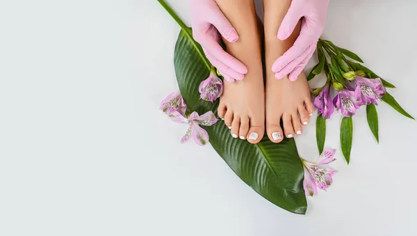 Beautiful perfect female skin legs feet top view with tropical flowers and green palm leaf. Nail polish, care and clean, spa pedicure treatment in white. Concept on background isolated. Copy space