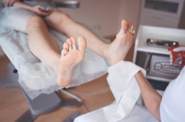 Ayak bileme ekipmanları kullanan ve ayak pedikürü için işlem cilası yapan pediatrist doktor. Beyaz eldivenli Podoloji Güzellik Uzmanı müşteri bacaklarını nasırdan ve mısırdan profesyonel aletlerle temizliyor.