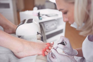 Pedikürcü, beyaz tırnak cilası sürüyor. Profesyonel tıbbi pedikür prosedürü. SPA salonunda ayak tedavisi. Ayak hastalıkları kliniği. Güzellik uzmanı beyaz eldivenlerle ellerini uzattı.