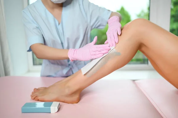 Woman with long tanned perfect legs and smooth skin having wax stripe depilation hair removal procedure on legs in beauty salon. Beautician in blue robe, pink gloves. Body care, epilation spa concept.