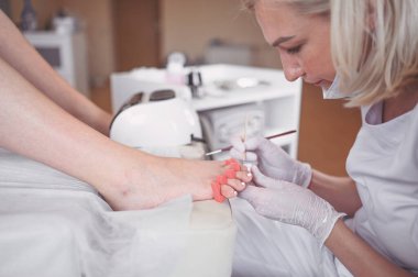 Pedikürcü, beyaz tırnak cilası sürüyor. Profesyonel tıbbi pedikür prosedürü. SPA salonunda ayak tedavisi. Ayak hastalıkları kliniği. Güzellik uzmanı beyaz eldivenlerle ellerini uzattı.
