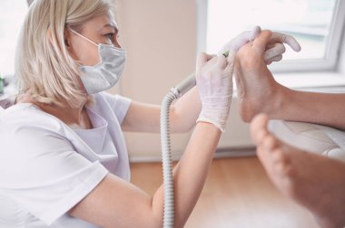Ayak bileme ekipmanları kullanan ve ayak pedikürü için işlem cilası yapan pediatrist doktor. Beyaz eldivenli Podoloji Güzellik Uzmanı müşteri bacaklarını nasırdan ve mısırdan profesyonel aletlerle temizliyor.